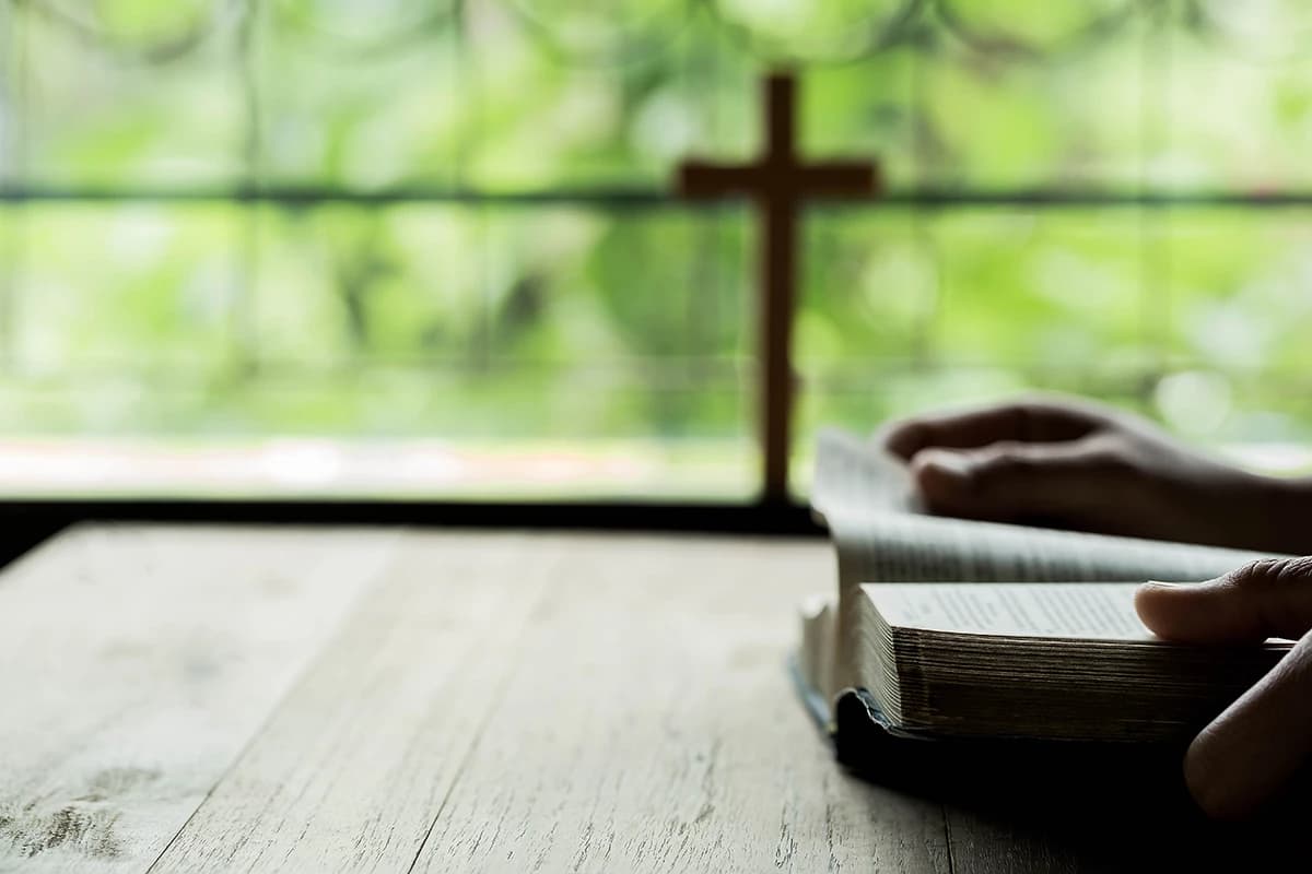 A person reads a Bible by a window with a wooden cross in the background symbolizing quiet reflection and the foundation of Bible study.