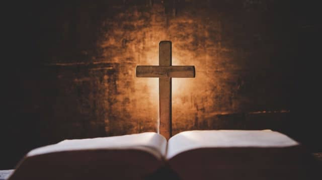 An open Bible with a wooden cross standing behind it, illuminated by soft light.